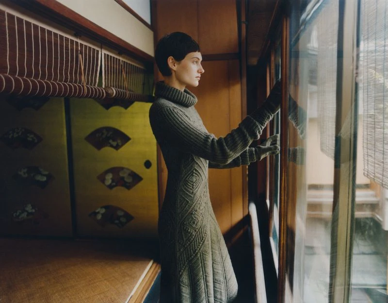 Woman in a gray dress gazes out a window in a traditionally styled Japanese interior with wooden walls and a decorative gold screen.