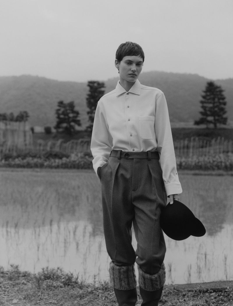 Monochrome fashion shot of a woman in a white shirt and trousers holding a hat, set against a backdrop of a field and mountains.