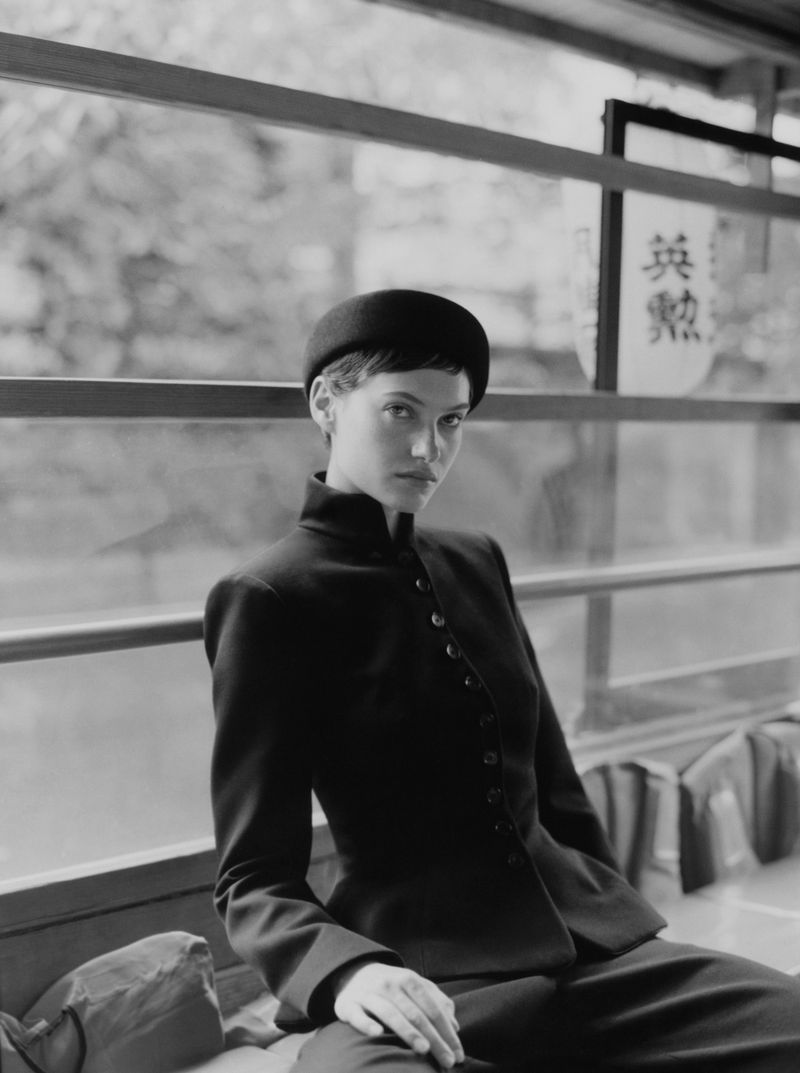 Monochrome portrait of a stylish woman in a tailored suit and matching hat, seated near a window with blurred foliage.