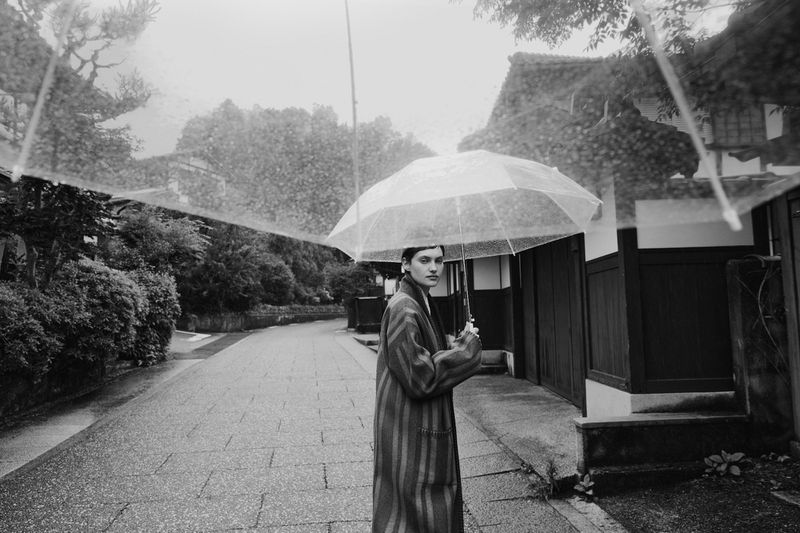 Black and white outdoor shot of a young adult wearing a striped coat, holding a transparent umbrella on a wet cobblestone street.
