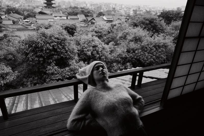 Monochrome photograph of a woman relaxing on a wooden surface with a backdrop of trees and rooftops beneath a cloudy sky.
