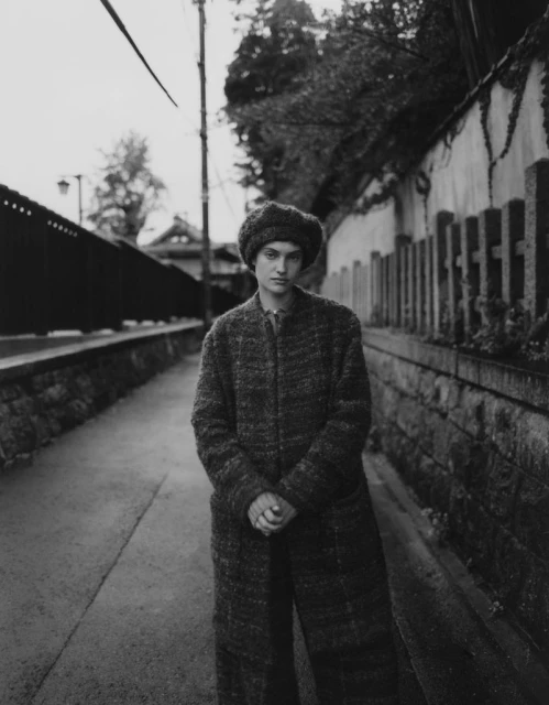 Monochrome portrait of a young woman in a coat and hat standing on a sidewalk in an urban setting with buildings and foliage.