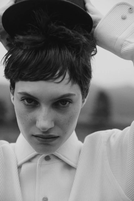 Black and white portrait of a young adult female wearing a collared shirt and hat, looking directly at the camera in an outdoor setting.