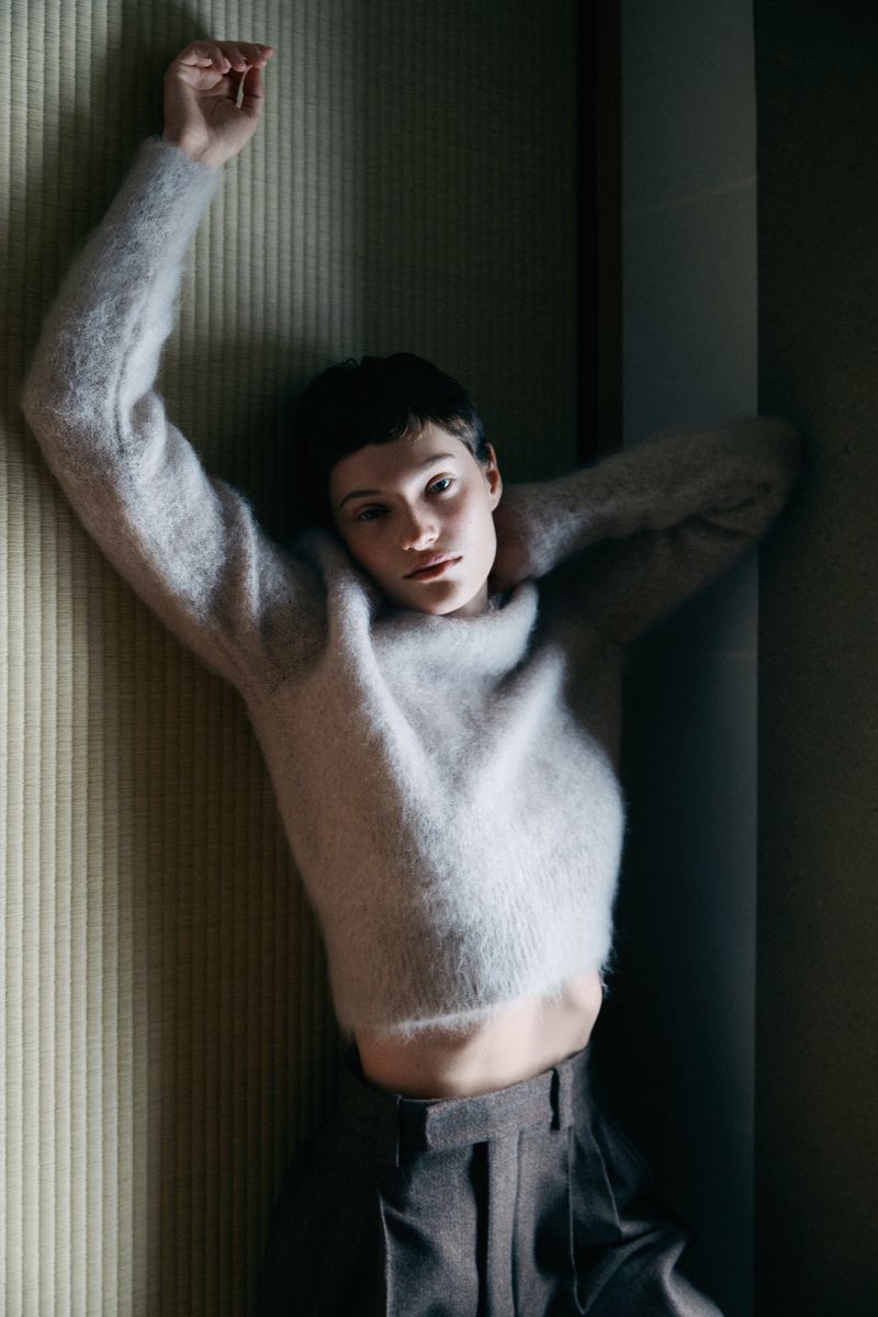 A young adult woman with short dark hair in a fuzzy grey sweater and grey pants poses in a corner in a dimly lit room.