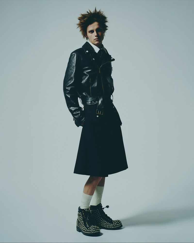 A male fashion model poses in a studio wearing a black leather jacket, dark skirt, white socks, and studded combat boots against a grey backdrop.