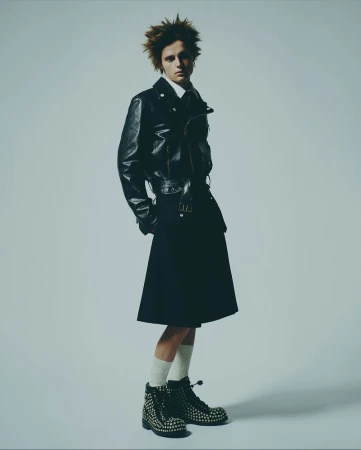 A male fashion model poses in a studio wearing a black leather jacket, dark skirt, white socks, and studded combat boots against a grey backdrop.