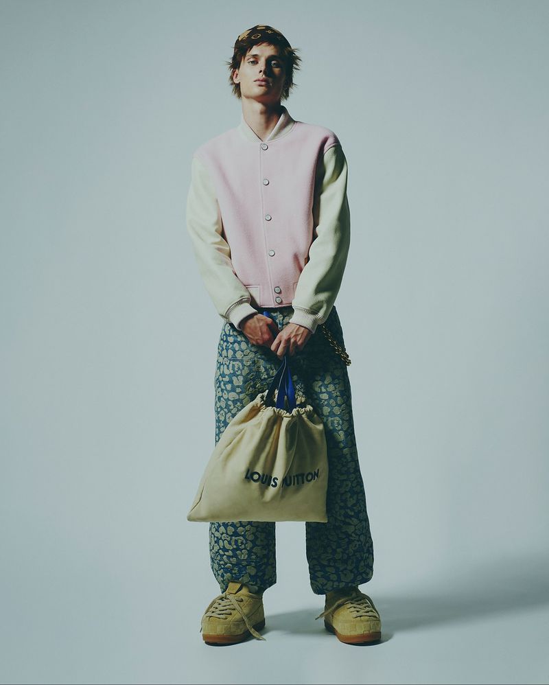 Young adult male fashion model poses with Louis Vuitton bag in a studio wearing a light pink jacket and patterned blue pants and beige sneakers.