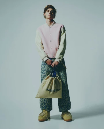 Young adult male fashion model poses with Louis Vuitton bag in a studio wearing a light pink jacket and patterned blue pants and beige sneakers.