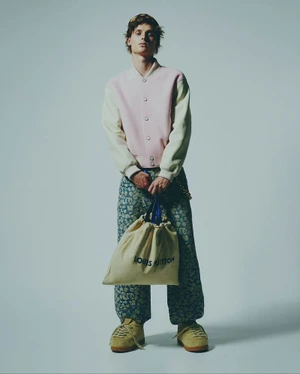 Male model wearing a pink and cream varsity jacket holding a Louis Vuitton dust bag against a grey background