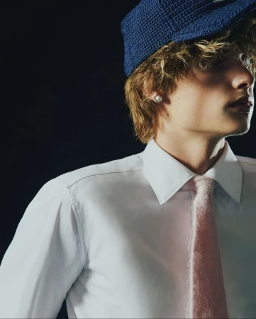 Close-up profile of a young male model wearing a white shirt, pink tie, and textured blue cap against a dark background.