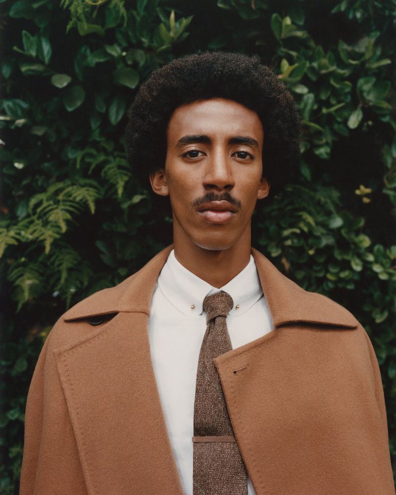 Portrait of a stylish adult man wearing a brown overcoat and white button-down shirt with tie, posing against a green leafy background.