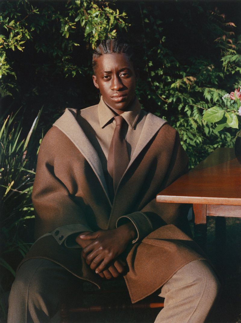 Portrait of a Black man wearing a brown coat and tie, seated outdoors against a lush green garden background.