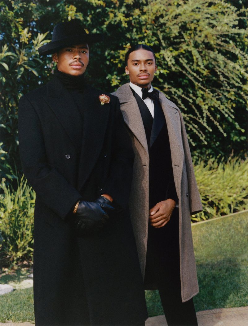 Two African American men pose outdoors, wearing overcoats and hats, exuding elegance and style against a natural green backdrop.