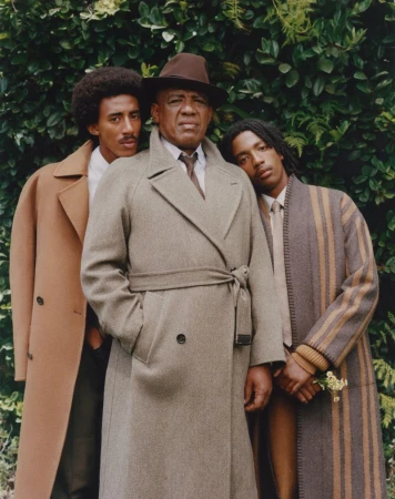 Three adult men stand outdoors wearing overcoats: the eldest wears a fedora while the two younger men stand either side of him.