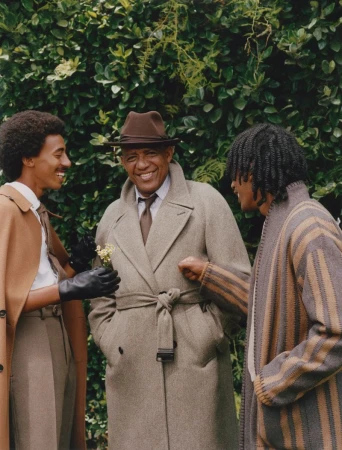 Three men of African descent are smiling outdoors; one holds flowers, wearing a hat and coat, with greenery in the background.