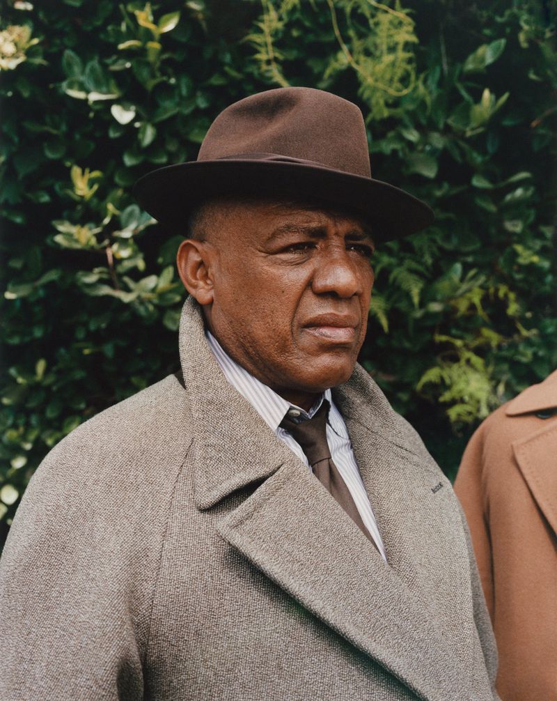 Headshot of an African-American man in a brown hat and gray coat, looking left, with green foliage visible in the background.