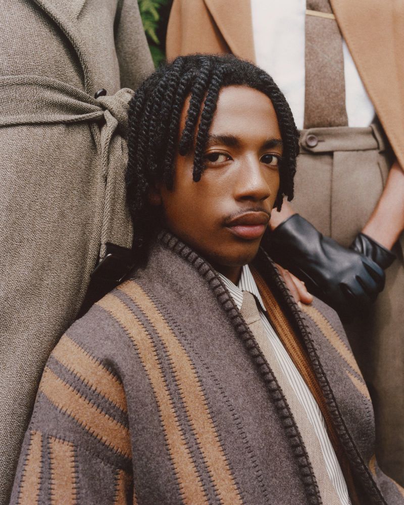 Close-up of a Black adult male with braided hair, wearing a blanket-style coat with a striped shirt and tie, surrounded by people in coats.