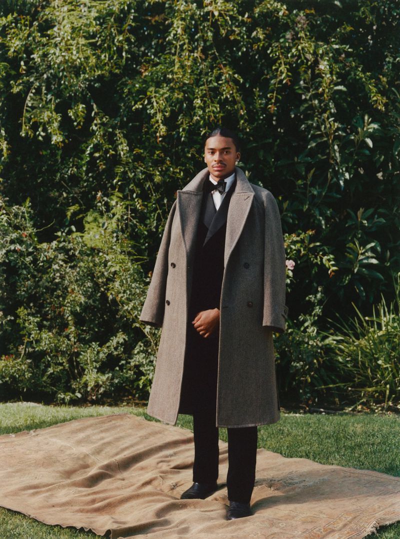 Man in a long coat stands in a garden setting, formally dressed with a bow tie, with a blanket at his feet, posing for a portrait.