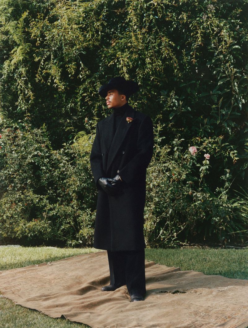 A stylish Black man in an all-black outfit, including a long coat and hat, stands confidently in a garden setting with lush greenery.