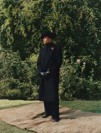 A stylish Black man in an all-black outfit, including a long coat and hat, stands confidently in a garden setting with lush greenery.