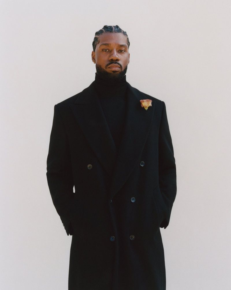 Portrait of a confident adult male of African descent wearing a black turtleneck sweater and a double-breasted wool coat in a studio.