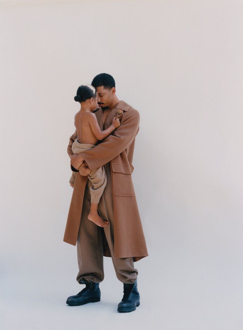 Father and child in neutral setting. African-American man holds young child, both wearing earth-toned clothing, posing against a plain backdrop.