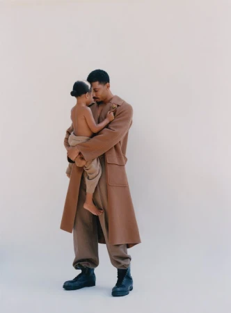 Father and child in neutral setting. African-American man holds young child, both wearing earth-toned clothing, posing against a plain backdrop.