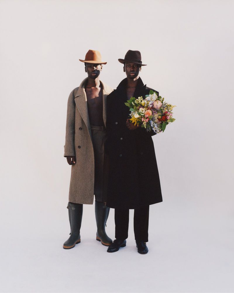Two dark-skinned adults in outerwear and hats stand in a neutral studio setting, one holding a colorful bouquet of flowers.