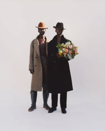 Two dark-skinned adults in outerwear and hats stand in a neutral studio setting, one holding a colorful bouquet of flowers.