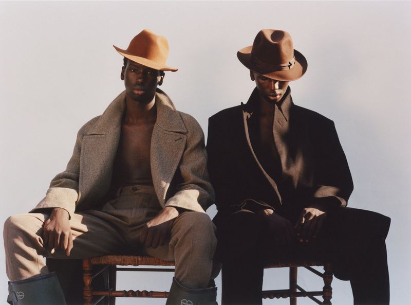 Two Black men sit side-by-side on wood chairs in a studio. They wear cowboy hats and overcoats in a fashion shot.