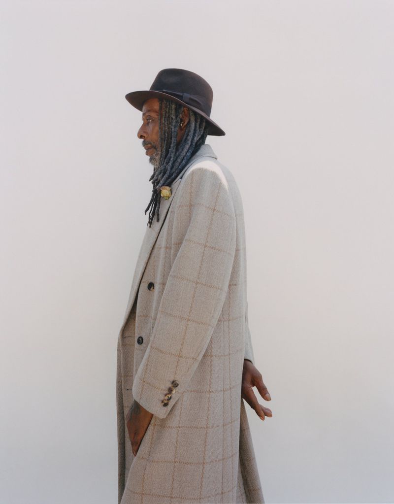 Profile portrait of a stylish Black man with dreadlocks wearing a hat and plaid overcoat, hands clasped, against a plain light background.