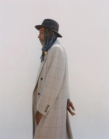 Profile portrait of a stylish Black man with dreadlocks wearing a hat and plaid overcoat, hands clasped, against a plain light background.