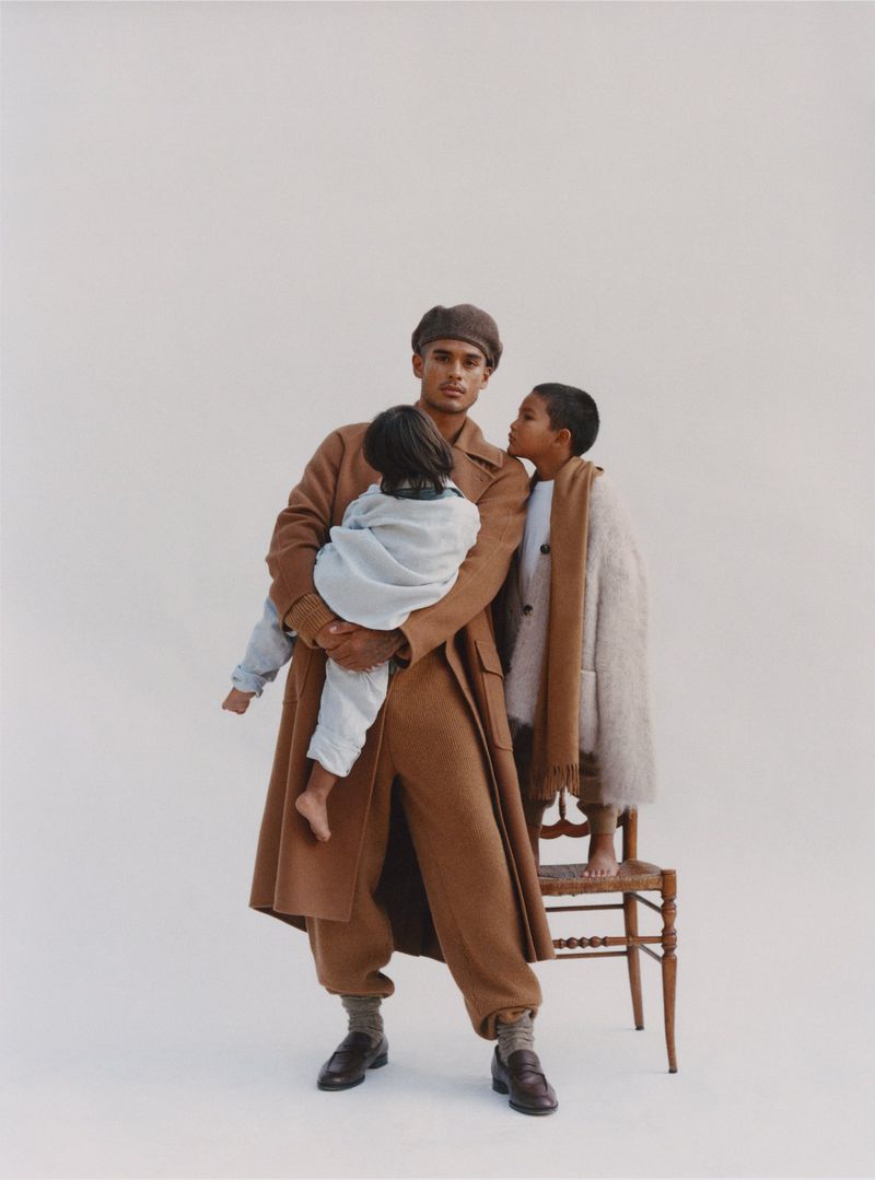 A father in a brown coat and matching pants holds his child while another child stands next to a wooden chair in a studio setting.