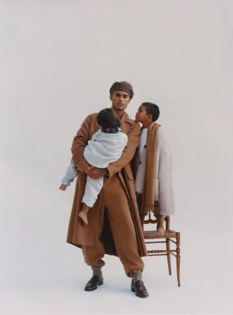 A father in a brown coat and matching pants holds his child while another child stands next to a wooden chair in a studio setting.