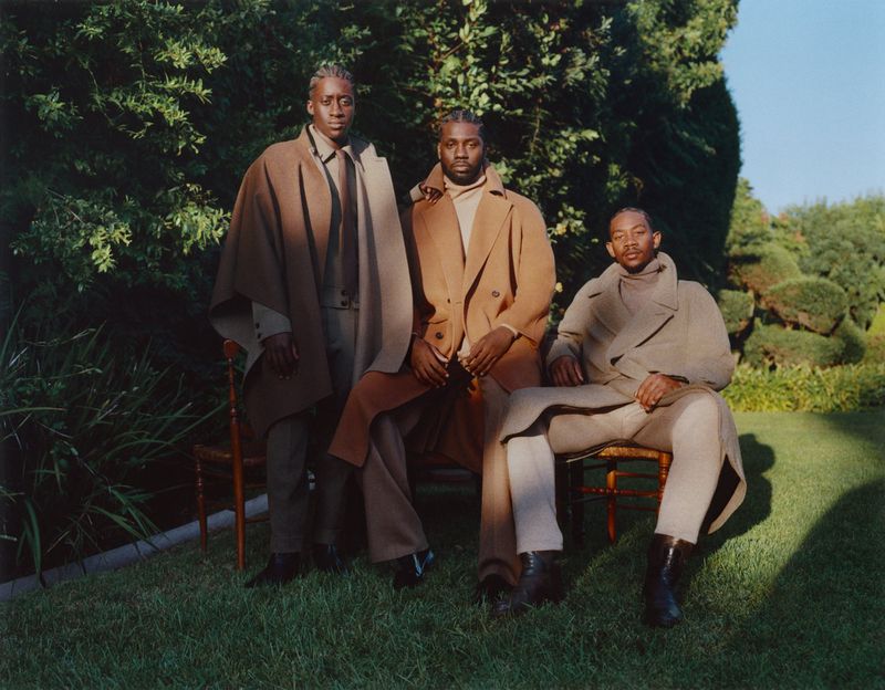 Three African-American men are sitting in a yard wearing coats and turtlenecks. They are in a natural setting with grass and trees.