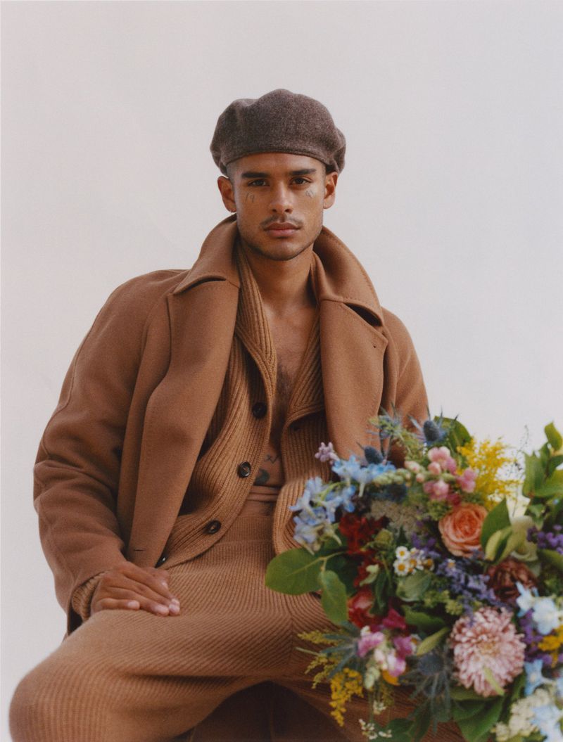 Portrait of a stylish man wearing a brown beret, coat, and matching knitwear sitting next to a vibrant bouquet of flowers in a studio.