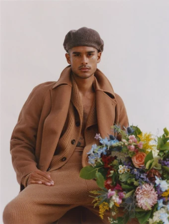 Portrait of a stylish man wearing a brown beret, coat, and matching knitwear sitting next to a vibrant bouquet of flowers in a studio.