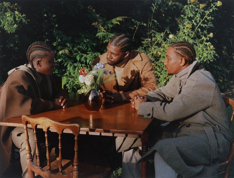 Three adult males sitting at a wooden table outdoors surrounded by green plants, wearing coats and braids, with a vase of flowers.