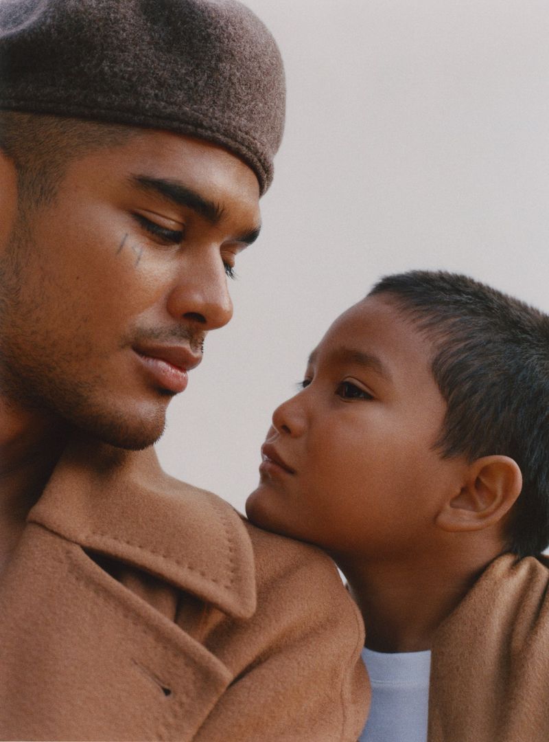 A young boy leans against an adult male in a studio portrait. The adult wears a beret and camel overcoat. Neutral background, medium shot.