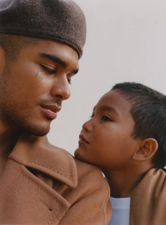 A young boy leans against an adult male in a studio portrait. The adult wears a beret and camel overcoat. Neutral background, medium shot.