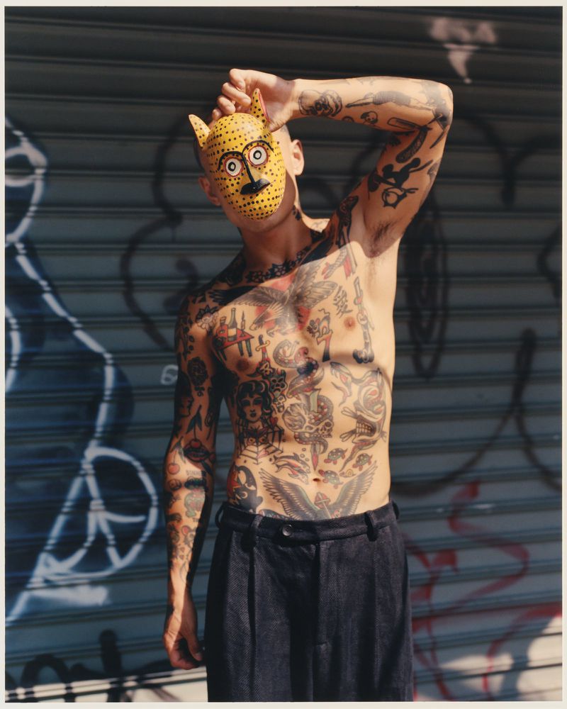 Tattooed man poses with a yellow leopard mask in urban setting. Graffiti wall provides backdrop. Street style and body art.