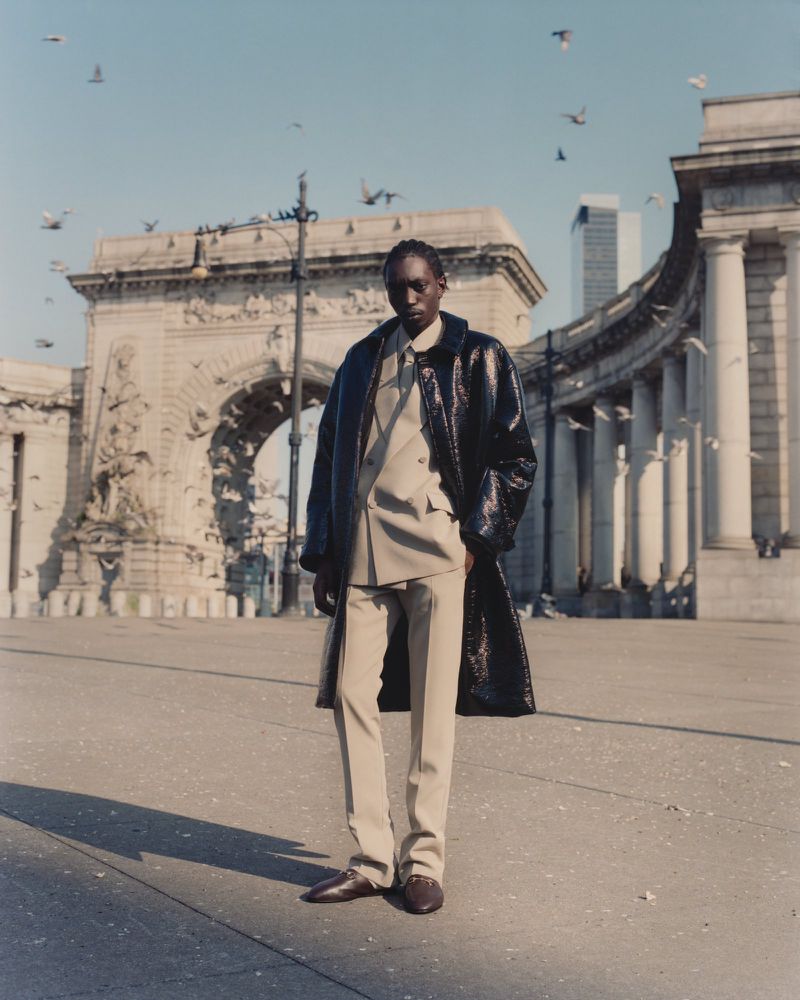 An adult Black man models a beige suit and dark coat outdoors in front of an archway and birds in flight on a sunny day.
