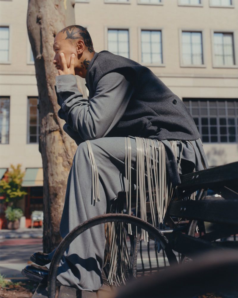 Man in grey suit sits outdoors next to a tree and vintage bicycle on a sunny day in front of a building with multi-paned windows.