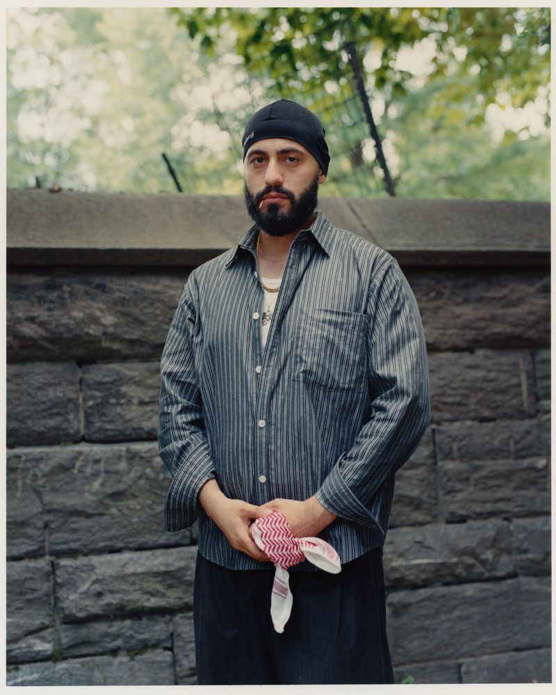 Portrait of adult male wearing a black head covering, striped shirt, and gold chain necklace, holding a plush rabbit toy against a stone wall.