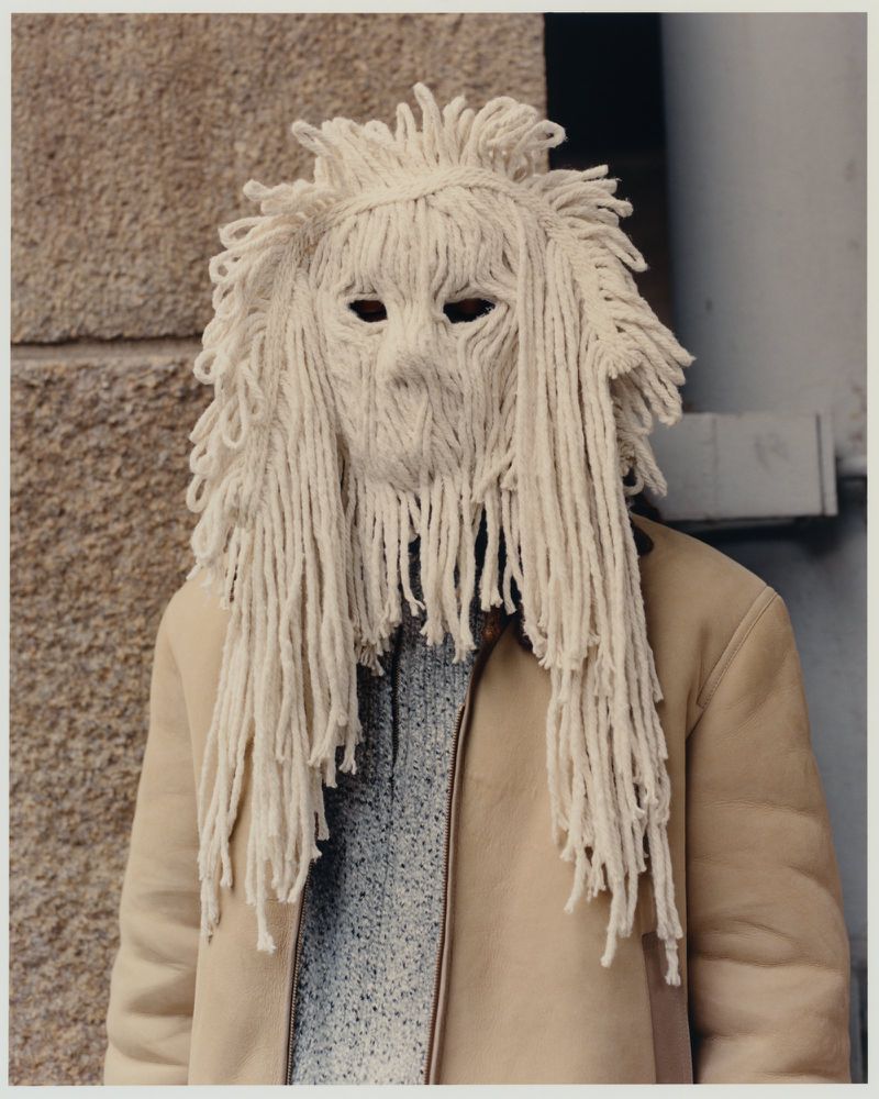 Person wearing a mop-like white mask and beige coat against a textured wall, creating an eerie, unsettling, and intriguing visual.