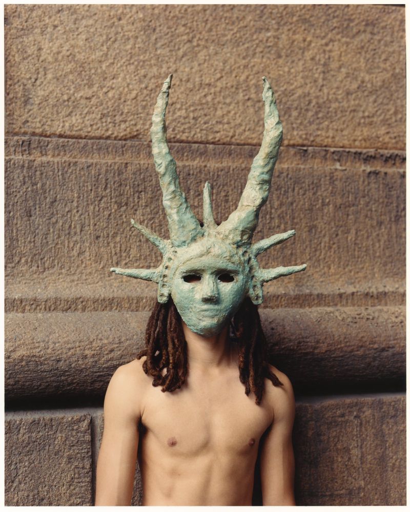 Man wearing Statue of Liberty mask stands against weathered stone wall in medium portrait. He has dreadlocks and is shirtless.