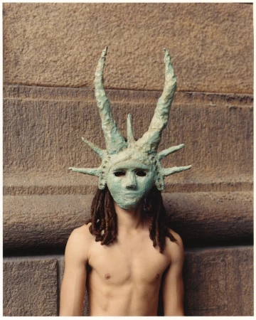 Man wearing Statue of Liberty mask stands against weathered stone wall in medium portrait. He has dreadlocks and is shirtless.