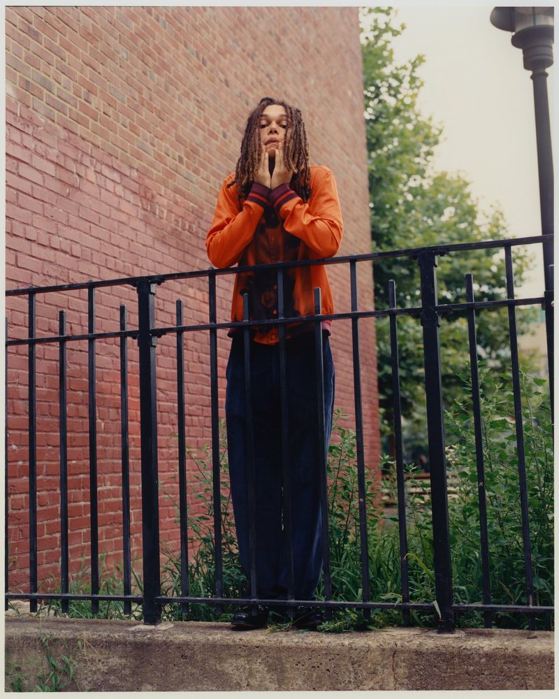 Young woman with dreadlocks, wearing an orange jacket and dark pants, stands behind a black metal fence in front of a brick building.