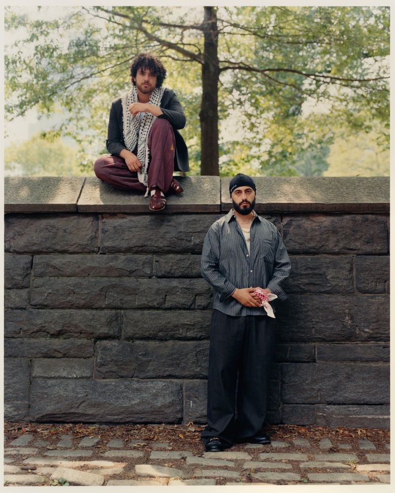 Two men posing outdoors against a dark stone wall with a tree behind them for an editorial feature in The Greatest Magazine.