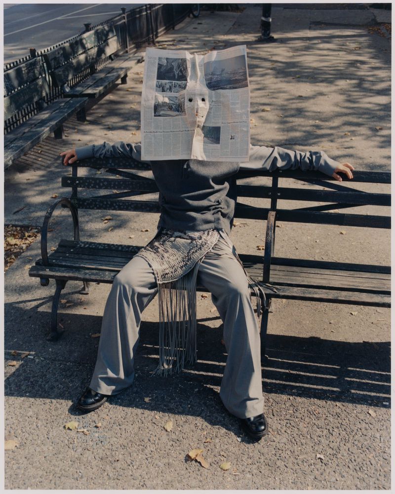 Person wearing a newspaper mask sitting on a park bench in sunlight. Figure is wearing a grey jacket and pants with black shoes.